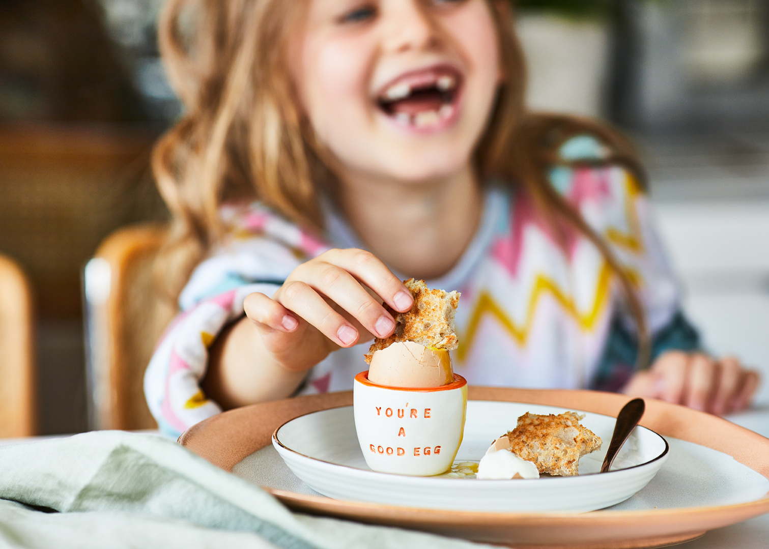 Still life product photography for online gift retailer Not on the High Street featuring a young girl eating a boiled egg from a personalised eggcup. Photography by London based food photographer Lol Johnson. Styling Nicki Bamford Bowes.