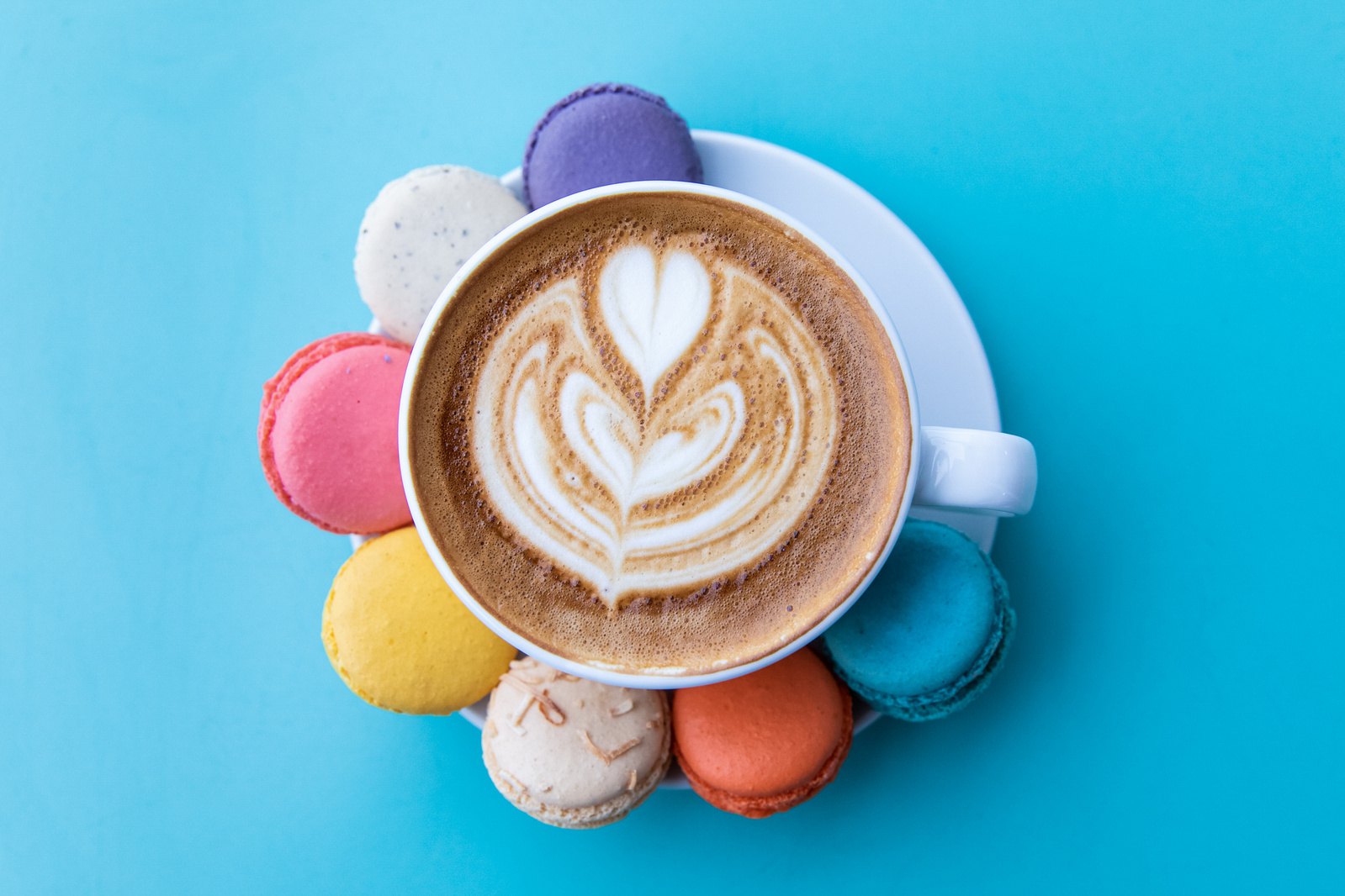 A latte with colorful macarons placed around the saucer on a blue background.