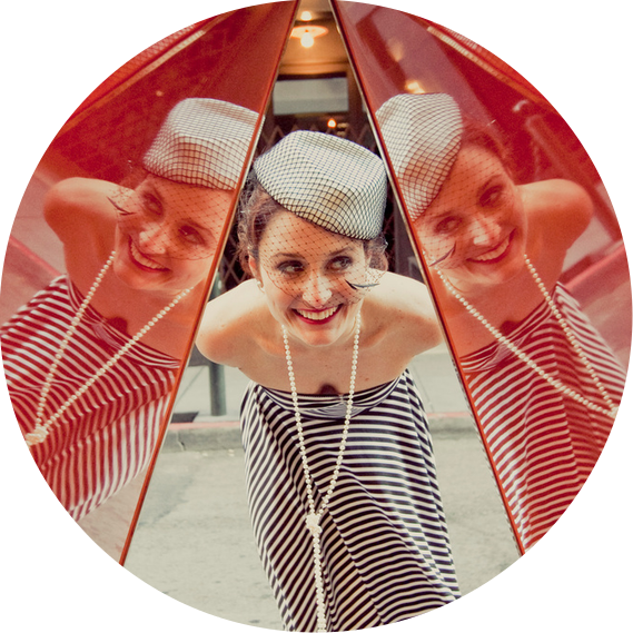 Woman in striped dress and small white hat bending over to see her reflection in a double mirror.