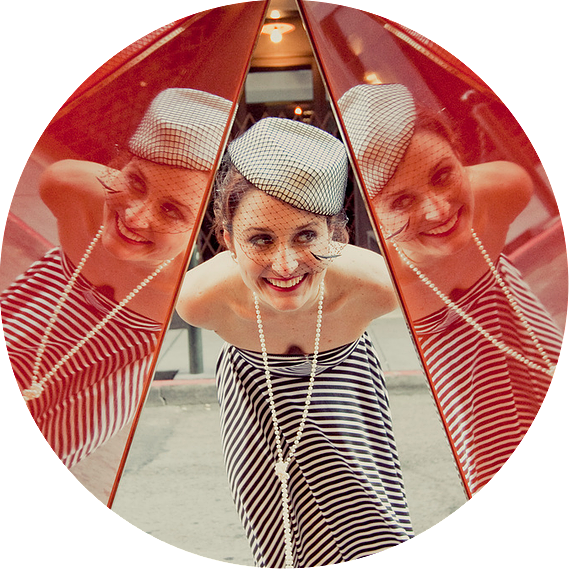 Woman in striped dress and small white hat bending over to see her reflection in a double mirror.