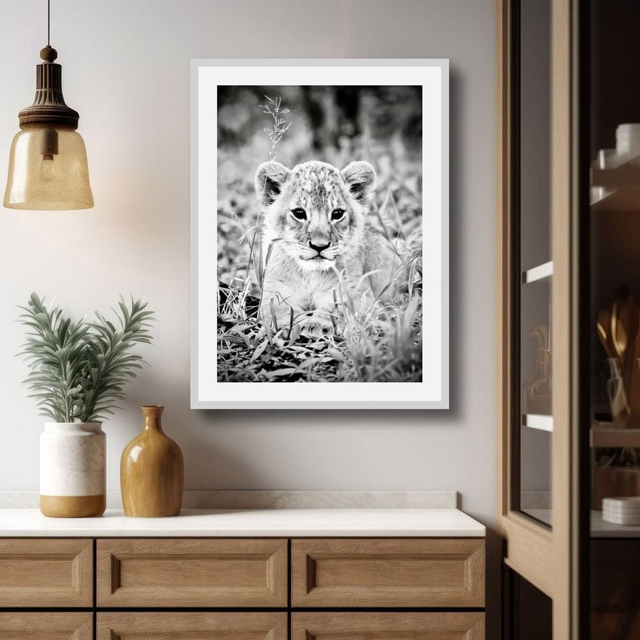Black and white fine art wildlife portrait of a young lion cub sitting in the grass, gazing directly into the camera with wide, expressive eyes. A powerful blend of innocence, curiosity, and early strength.