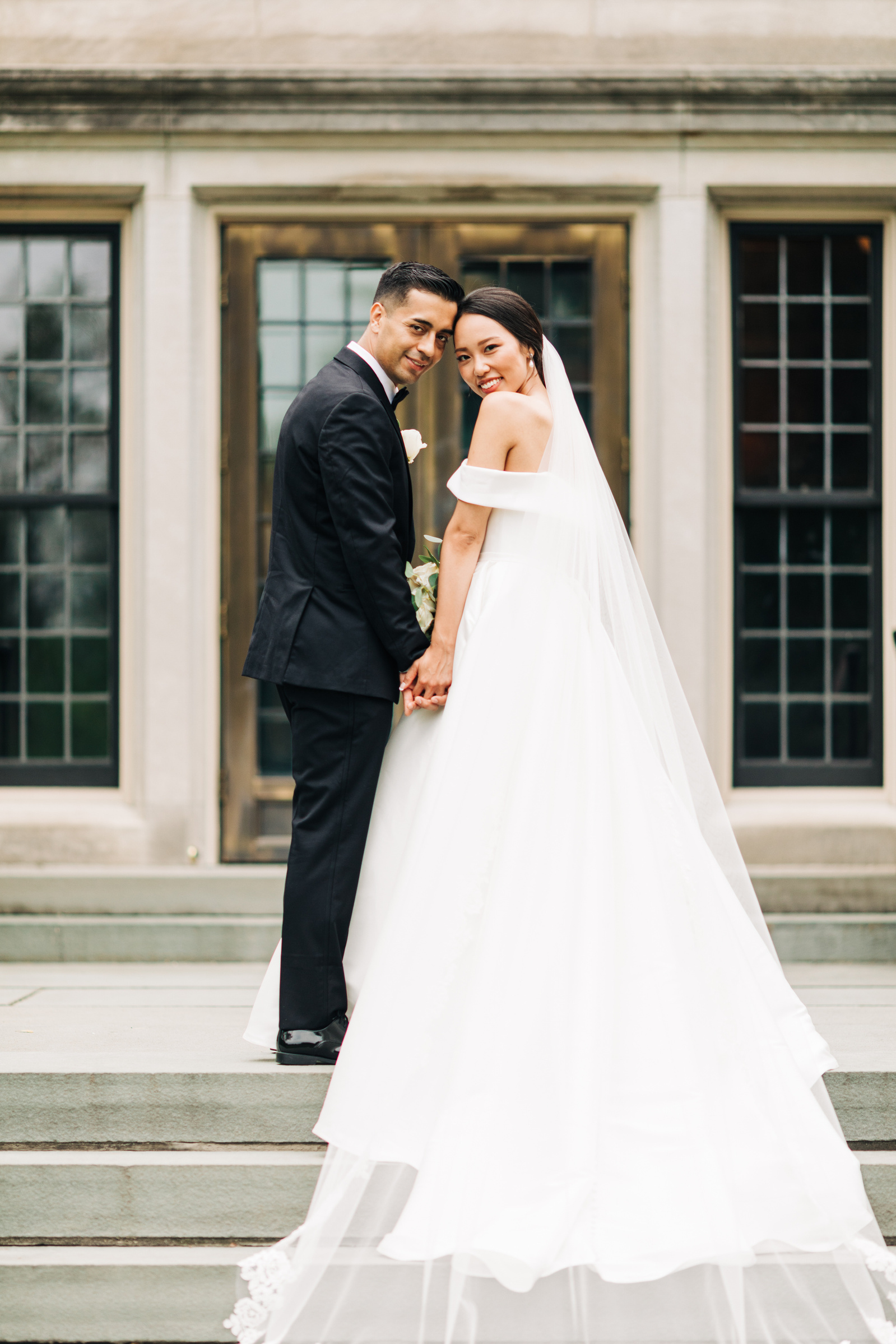 bride and groom hold hands standing on stairs and looking back at the camera as bride's veil is draped down the stairs