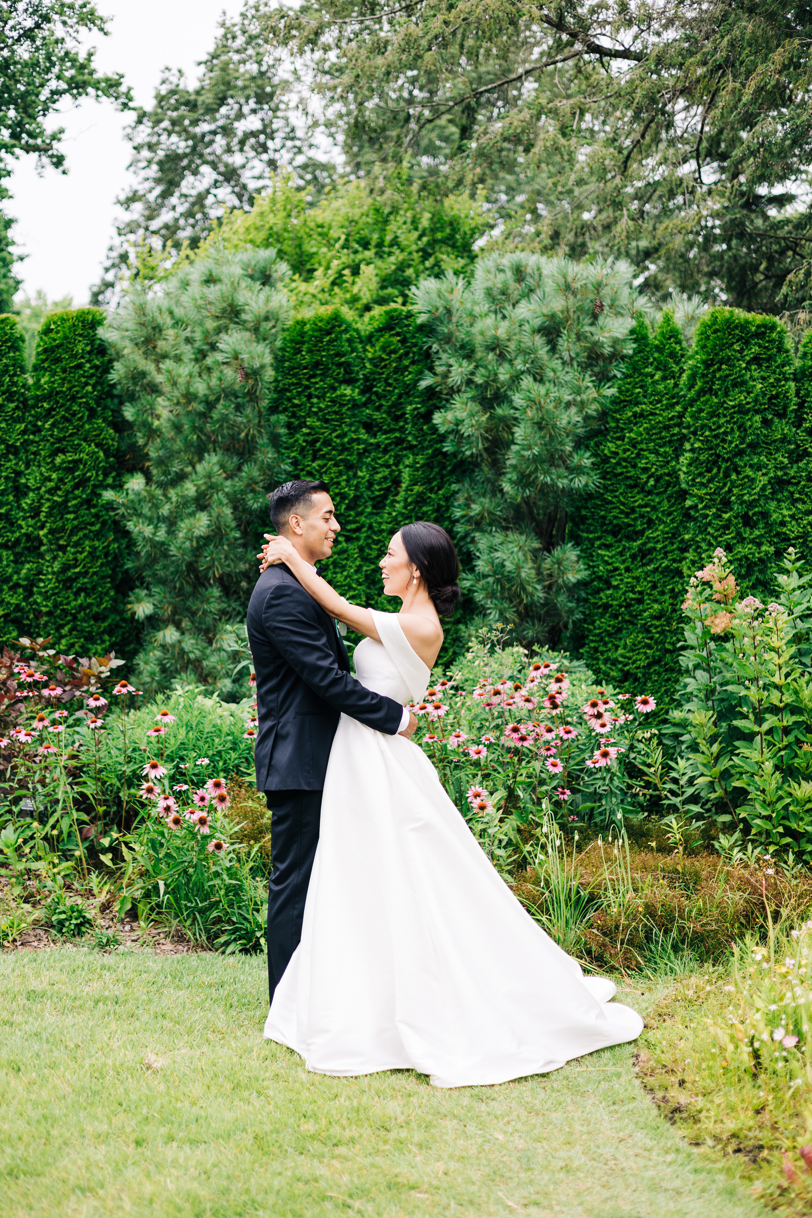 bride and groom standing in a garden and embracing during their wedding portraits with greenery and pink flowers in background
