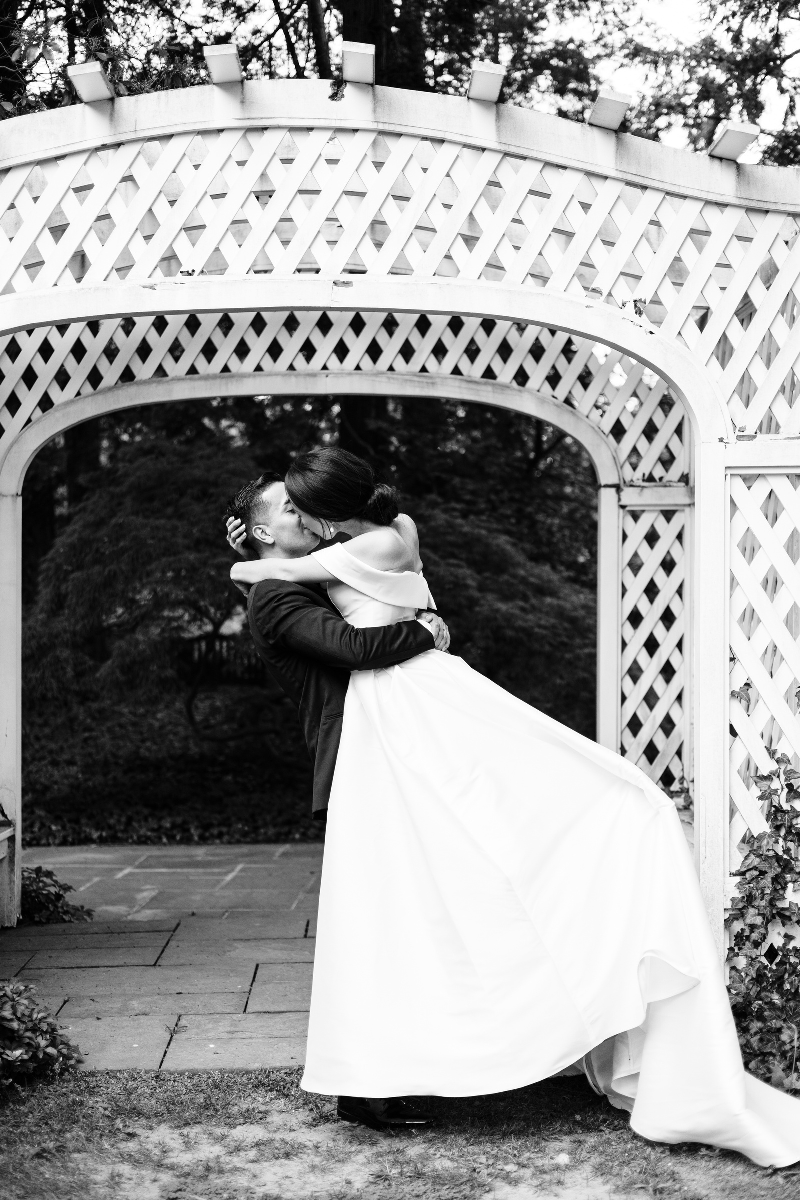 black and white photo of groom lifting up his bride and kissing her in front of a white garden arch