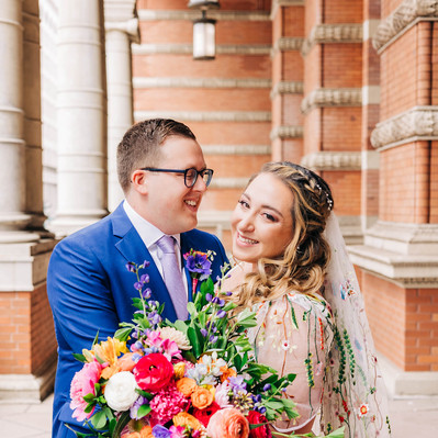 Bride and groom smiling on their wedding day. Groom is wearing a bright blue suit. Bride is holding a big colorful bouquet. Her wedding dress has colorful floral embroidery. 