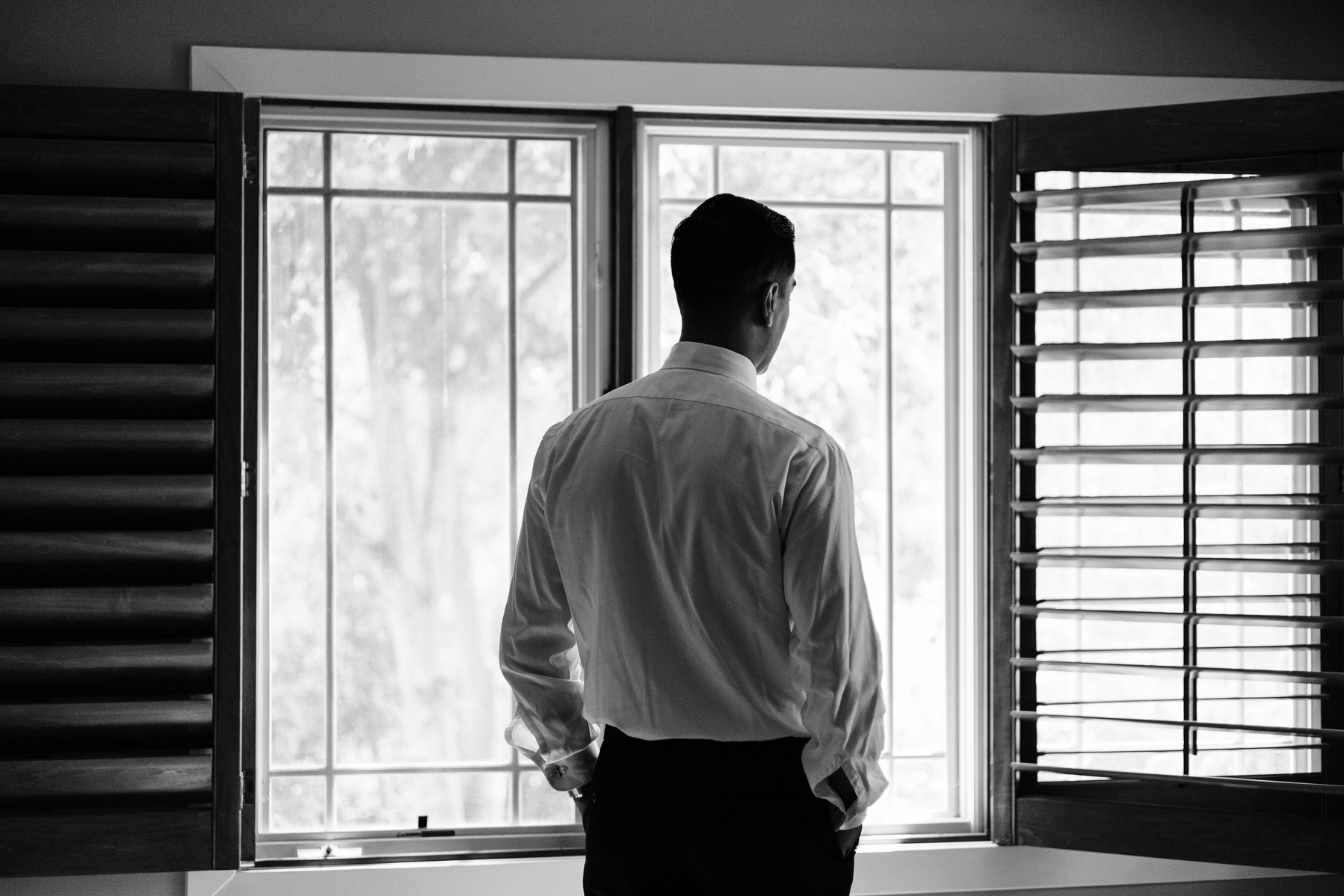 black and white photo of groom looking out the window while getting ready for his wedding