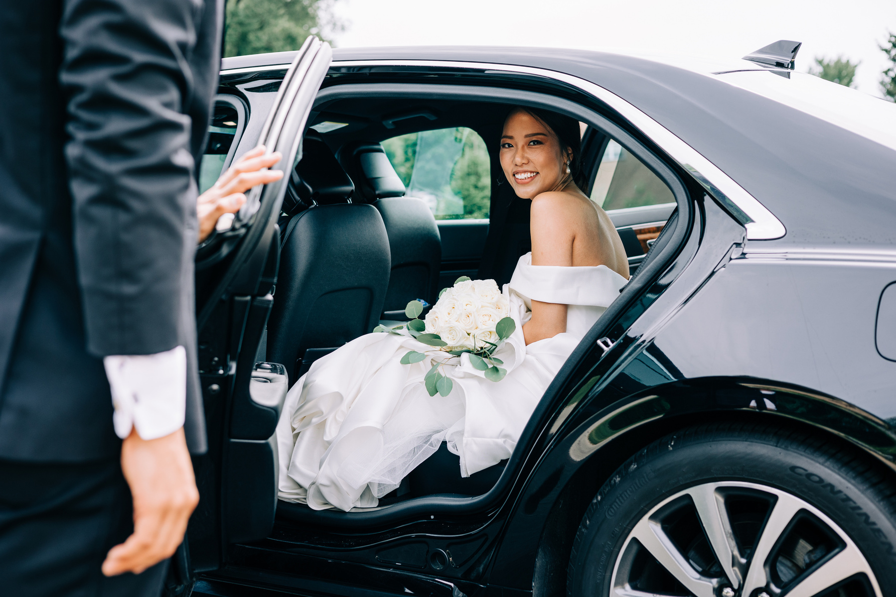 bride sits in a limo after their wedding ceremony as groom helps her in