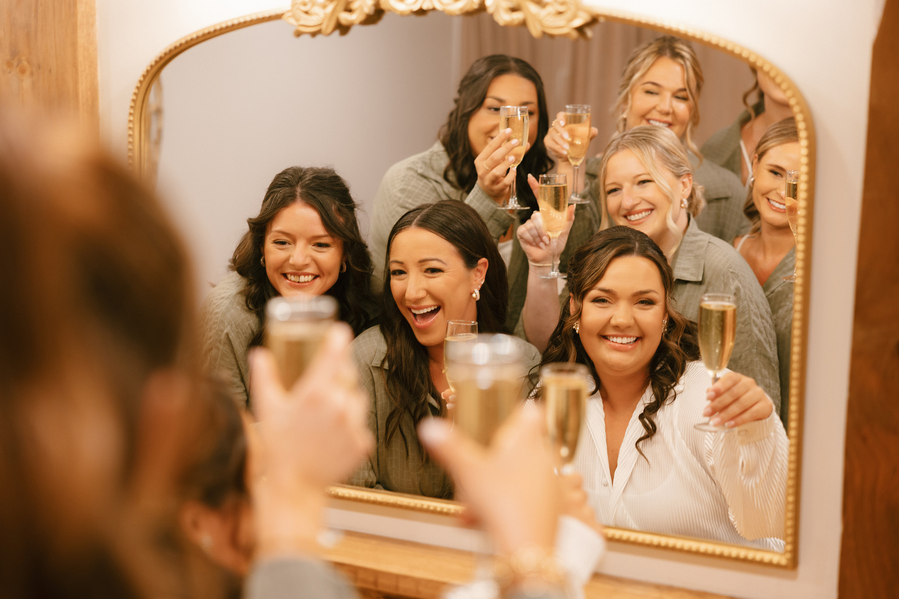 Bridesmaids are holding up champagne glasses and cheering in front of the mirror. 