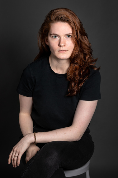 A white nonbinary human with long wavy red hair wears all black and sits on a stool while looking directly into the camera. 