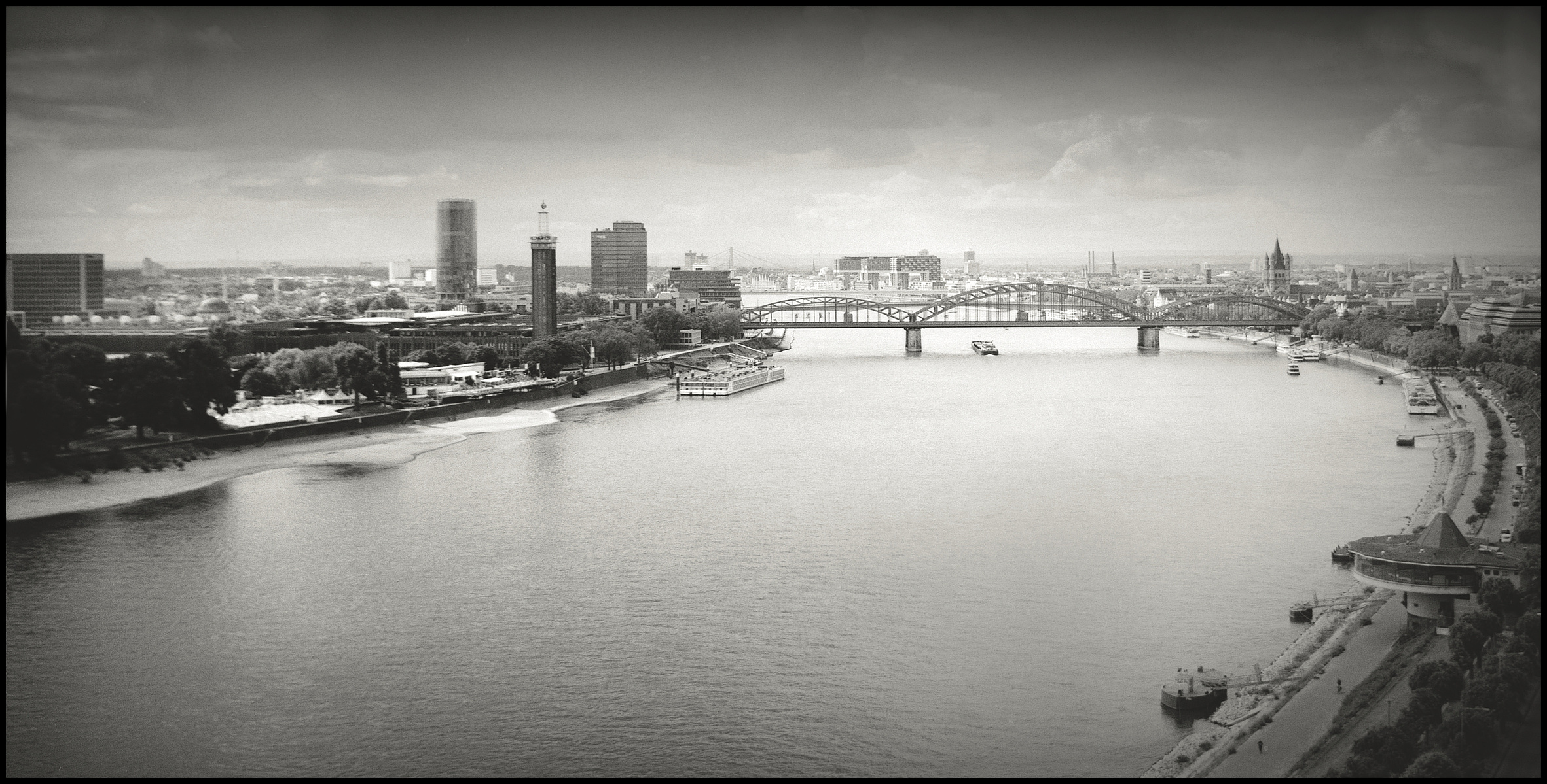 a black an white analog photography with a view over the river Rhine and the Hohenzollernbrücke. In the background of the photo are the "Kranhäuser"
