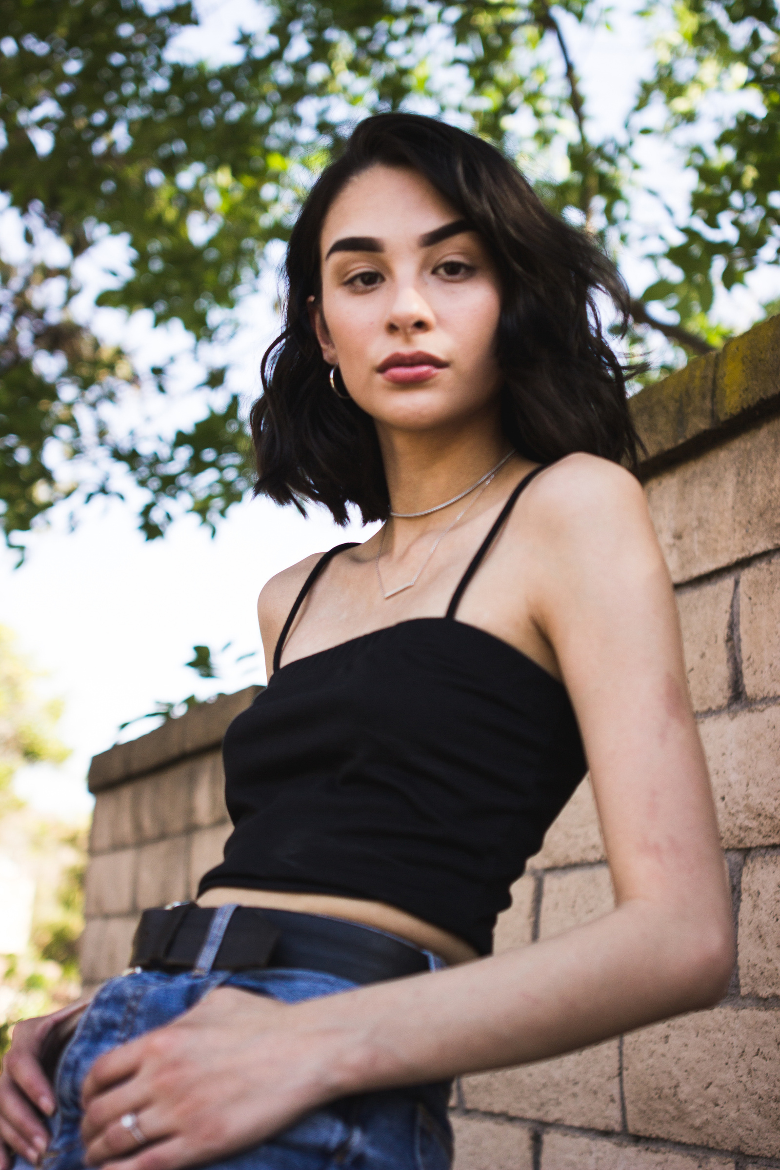 Woman wearing black top and jeans, standing in front of tree and brick wall