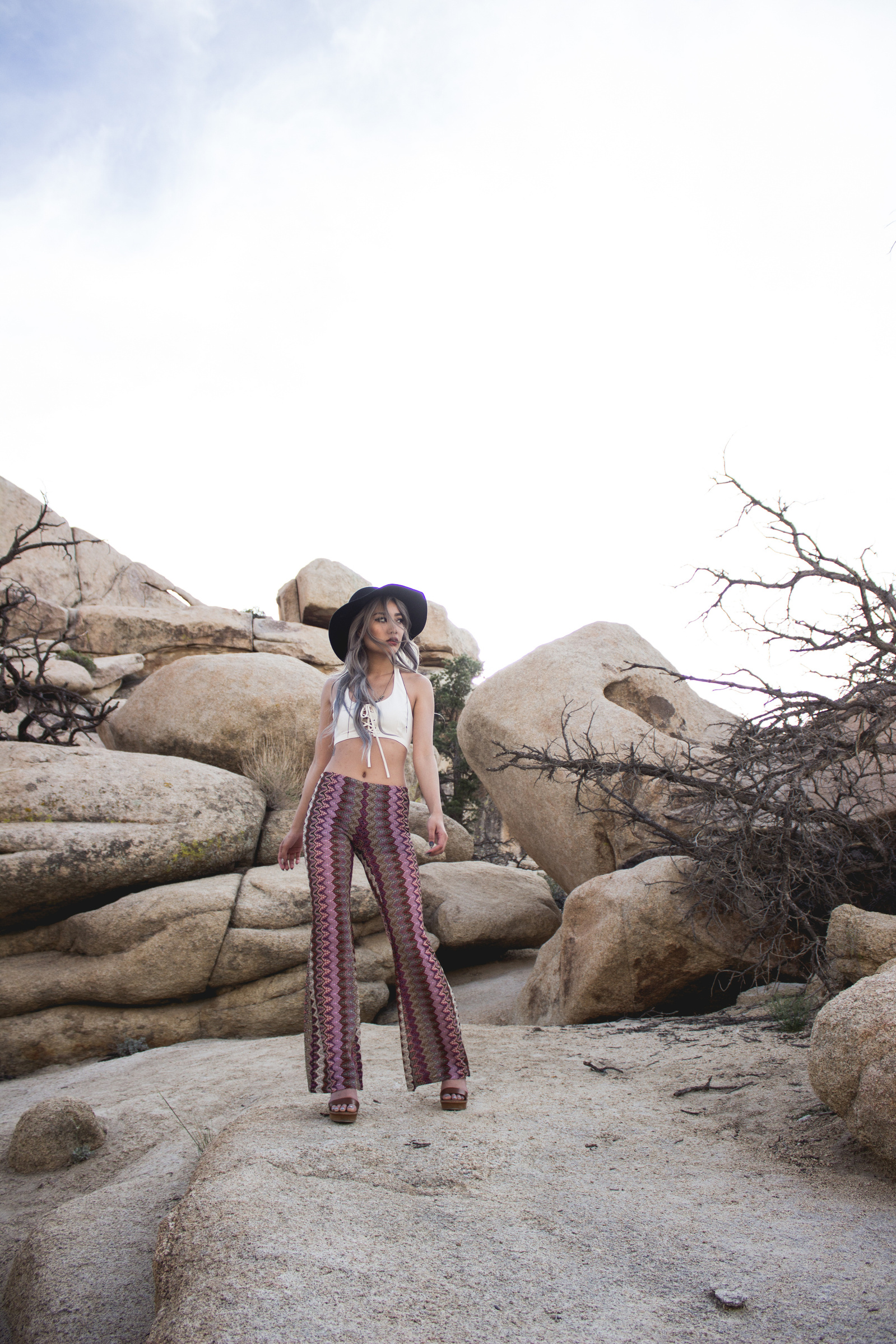 Woman wearing white top, black hat and flare pants, standing on rock formation with blue sky behind