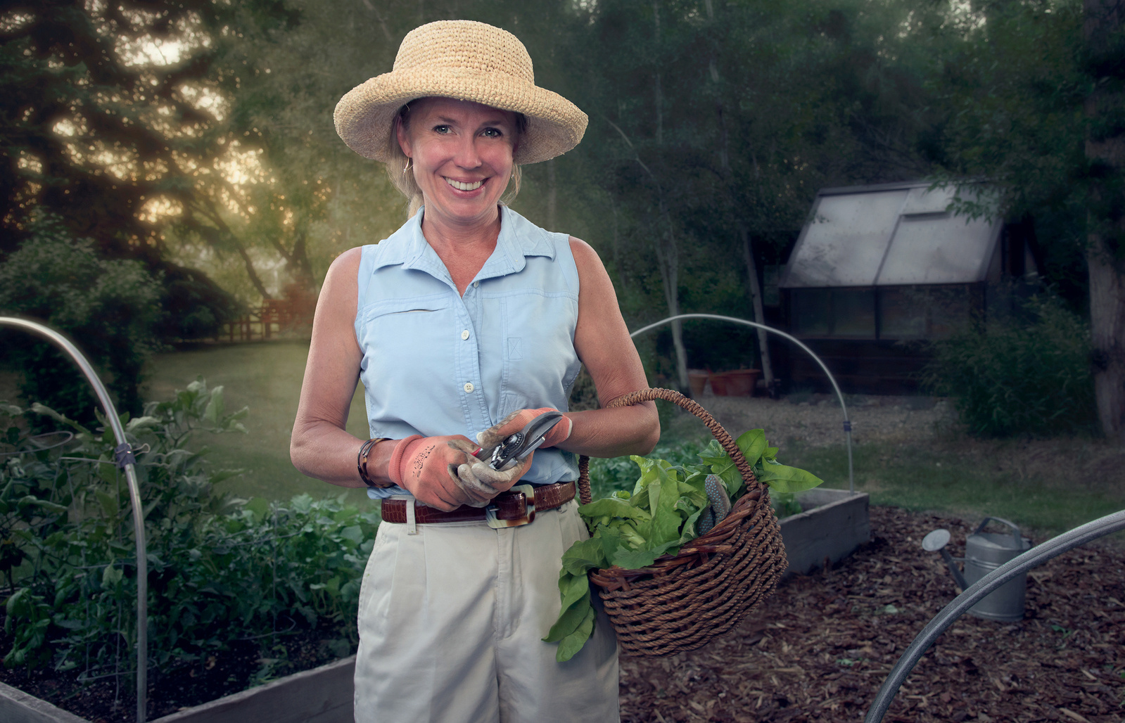 Woman in straw hat and blue shirt in her backyard garden, holding a basket of fresh-cut lettuce.