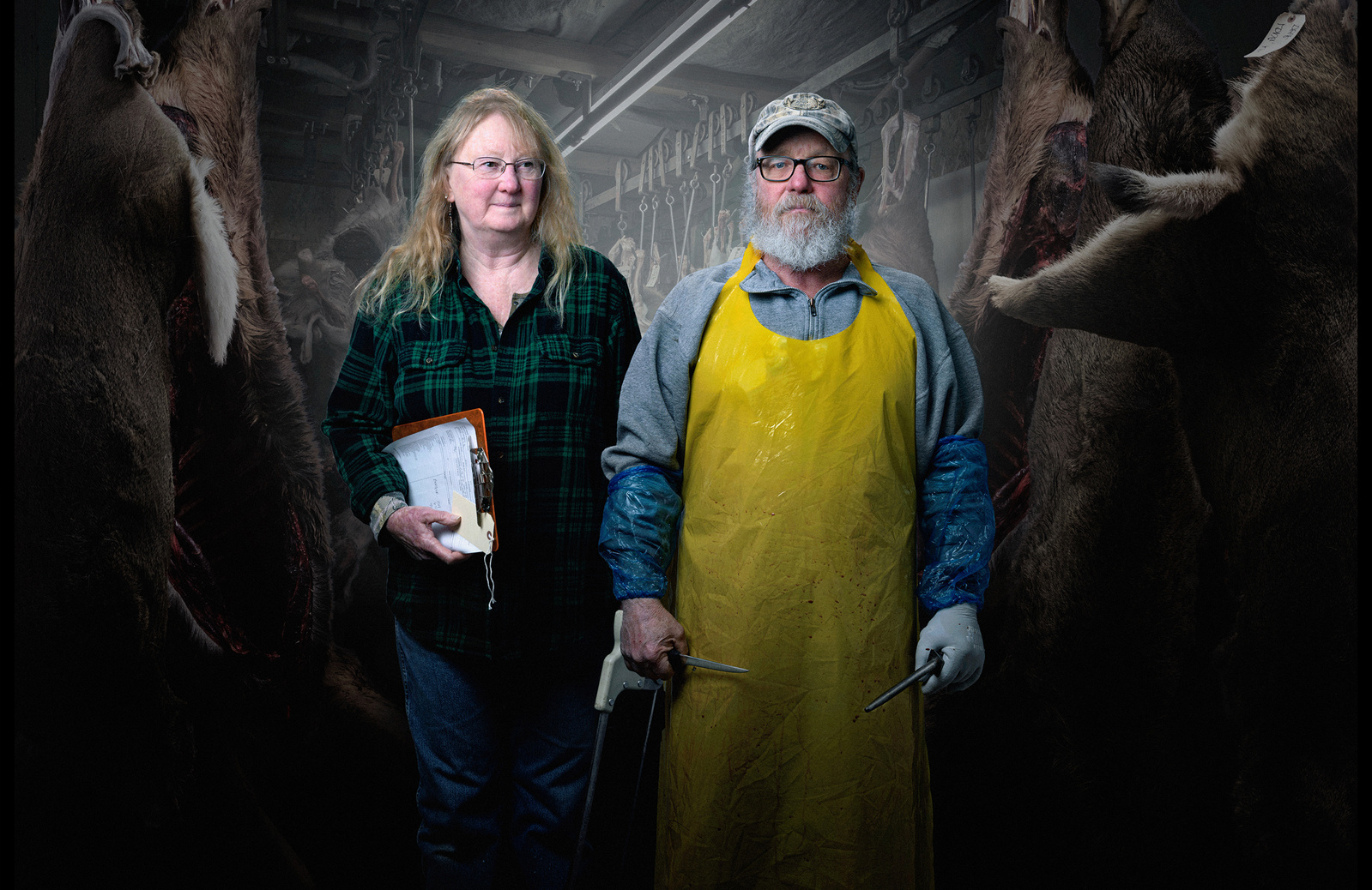 A portrait of Buzz and Patti Jones of Yellowstone Game Processing pose in front of elk and deer aging in a meat locker.