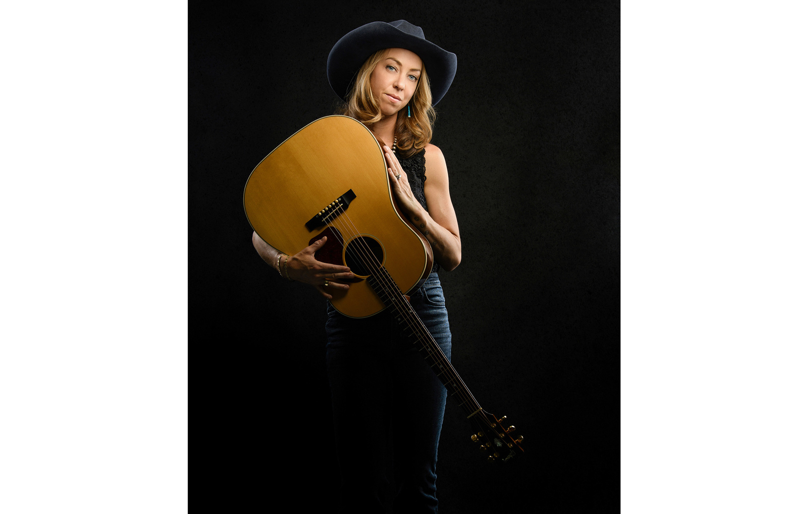 Studio portrait of a beautiful blonde-haired woman wearing a blue cowboy hat and cradling an acoustic guitar.