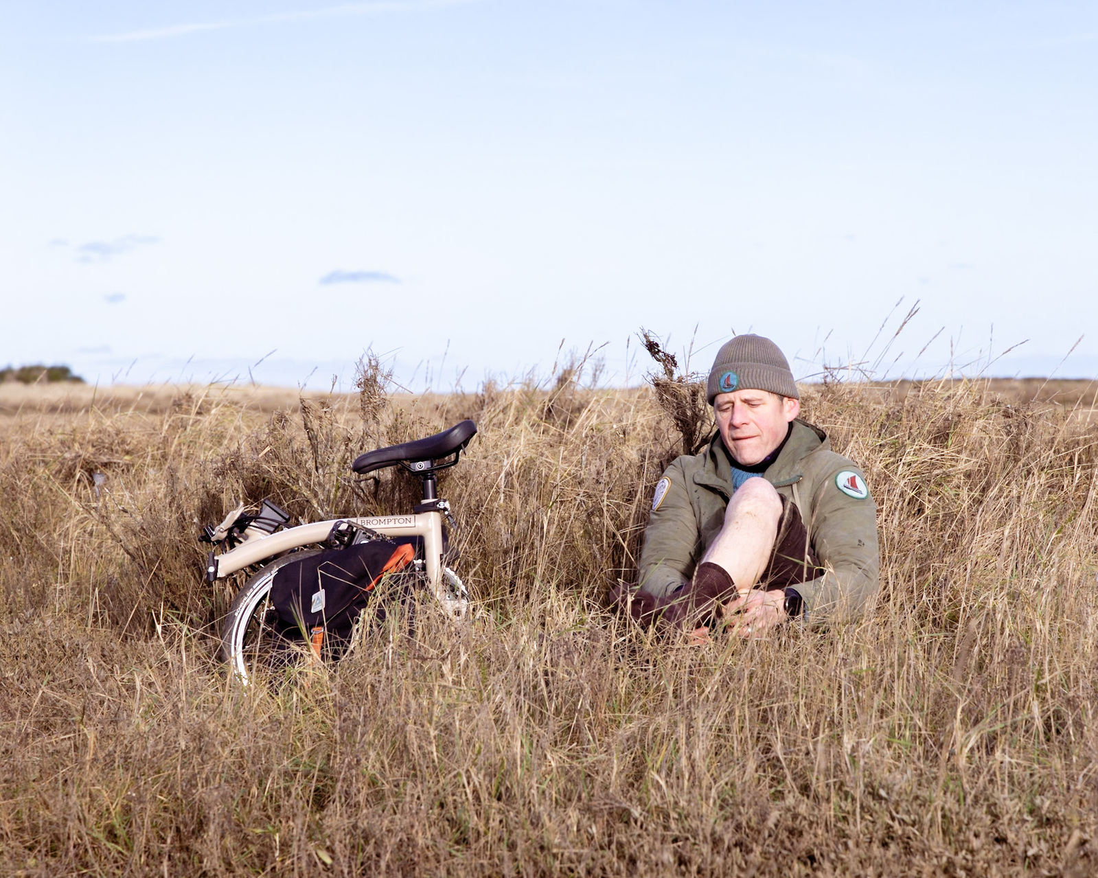 A man sat in a salt marsh, putting on dry socks.  His folded Brompton is next to him.
