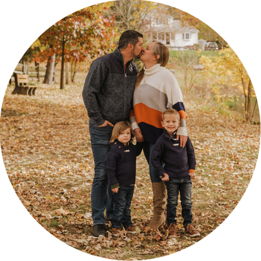 The Stephenson family stands together under a fall canopy of yellow trees. The parents kiss, while the two younger boys look towards the camera and smile. 

They are casually dressed in jeans and sweaters. 