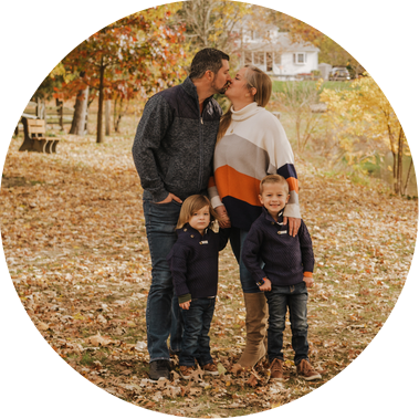 The Stephenson family stands together under a fall canopy of yellow trees. The parents kiss, while the two younger boys look towards the camera and smile. 

They are casually dressed in jeans and sweaters. 