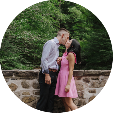 Anna & Jimmy share a kiss at Bowman's Wildflower Preserve during their proposal and engagement session.

Anna wears a bright pink dress and Jimmy wears a blue long sleeve with slacks. They are framed against a stone bridge and lush greenery behind them.
