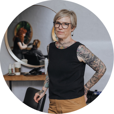 Heather poses with her salon chair in her original Halo Hair salon during her professional portrait session.

She is heavily tattooed on her arms, with close cropped hair and glasses. Behind her, a mirror reflects another employee styling a client.