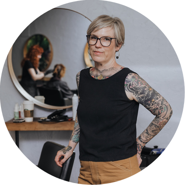 Heather poses with her salon chair in her original Halo Hair salon during her professional portrait session.

She is heavily tattooed on her arms, with close cropped hair and glasses. Behind her, a mirror reflects another employee styling a client.