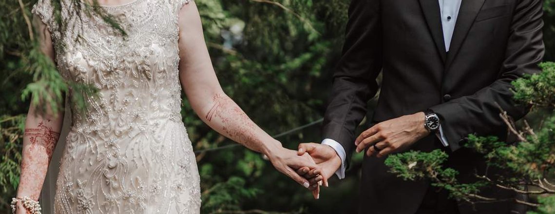 A close up, boho photograph of a newly married bride and groom holding hands in the woods. The bride is wearing a detailed ivory gown, with henna tattoos on her arms. The groom is wearing a black tuxedo with a fancy watch.  Lauxmont Farms, Lancaster, PA.