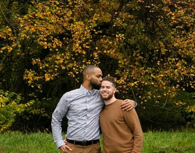 A gay couple stands in front of a background of fall foliage, enjoying their time together during their engagement session in Bellevue State Park, in Delaware.