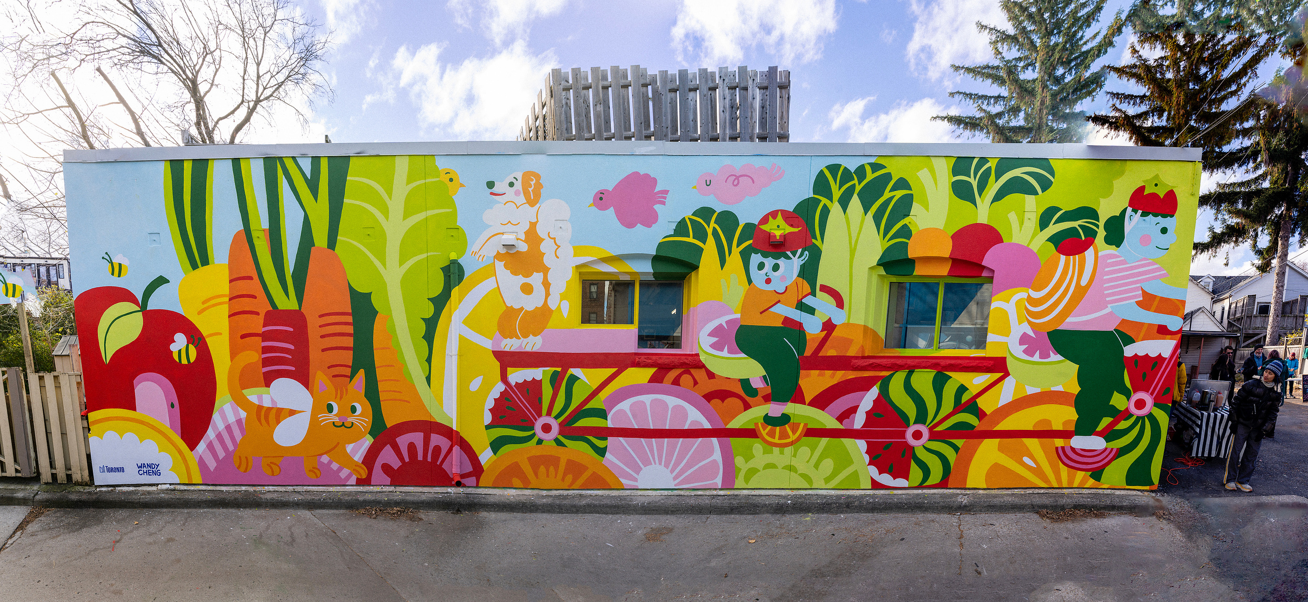 A public colour mural of a family biking on a long bike made of watermelon wheels with a citrus landscape, followed by a giant orange cat with a dog being surprised at a bird in a lettuce leaf.