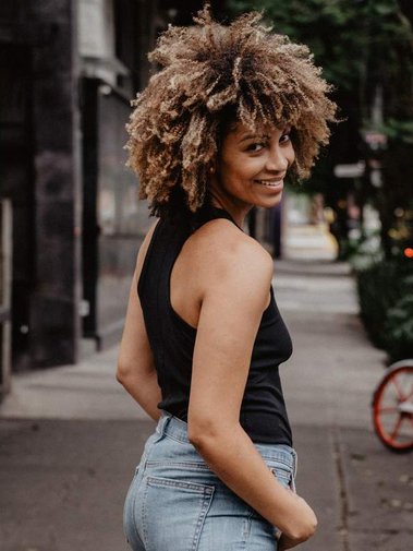 natural curly hair light skin black women in black tan and light jeans looking over shoulder and smiling walking down tree-lined street