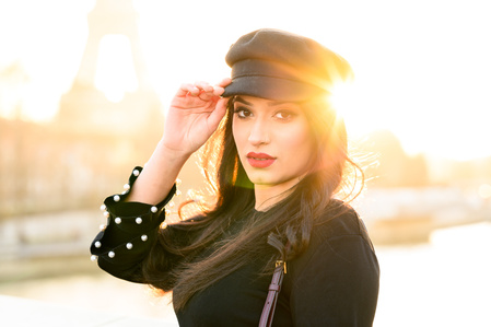 Photo prise à contre-jour en bord de Seine à proximité de la Tour Eiffel d&#x27;une jeune femme maintenant sa casquette avec la main droite