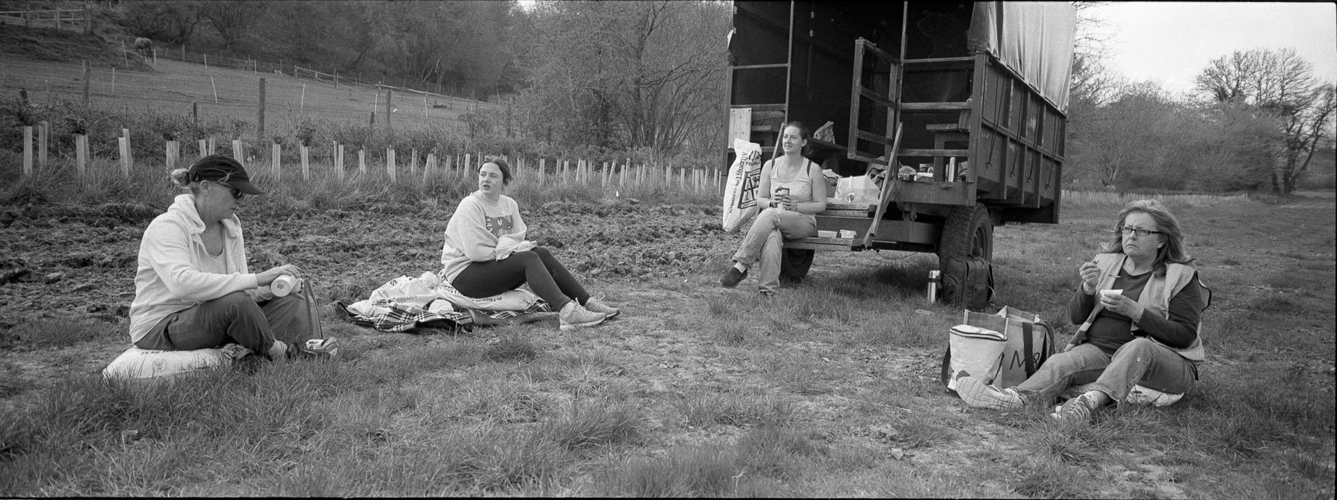Four female volunteers sit by a Shepherd hut on a lunch break in the Hampton Estate hop fields in Puttenham, Surrey. The black and white wide format photograph was made using a Hasseblad Xpan camera and Ilford HP5 Plus film.