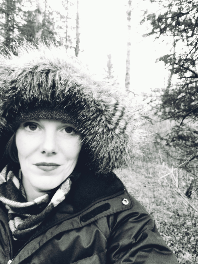 Picture of Torina Stark. Torina is wearing a thick black winter coat with fake fur framing her white face. The picture is in black and white set in the woods with balsam fir trees and trail behind.