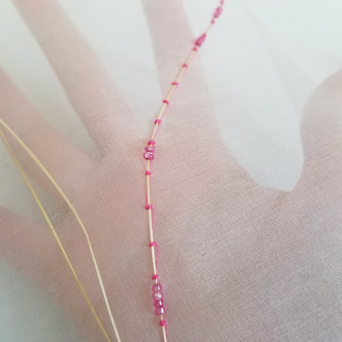 Organza fabric with my hand behind it so you can visualize the transparency. Three dried grass blades on top with one couched onto the fabric with thick pink thread and pink glass beads