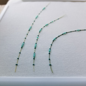Art: Three dried grass blades couched onto a white fabric background with thick green thread and seafoam green glass beads