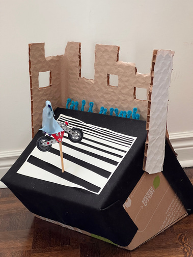 Three quarter view of a cardboard box sitting at an angle, with cardboard buildings attached at the back. A wooden dowel holds up a metal toy motorcycle, and blue human shaped pipe cleaners sit in front the buildings