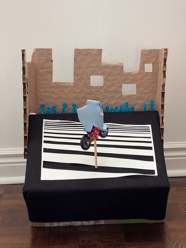 Front on view of a cardboard box sitting at an angle, with cardboard buildings attached at the back. A wooden dowel holds up a metal toy motorcycle, and blue human shaped pipe cleaners sit in front the buildings