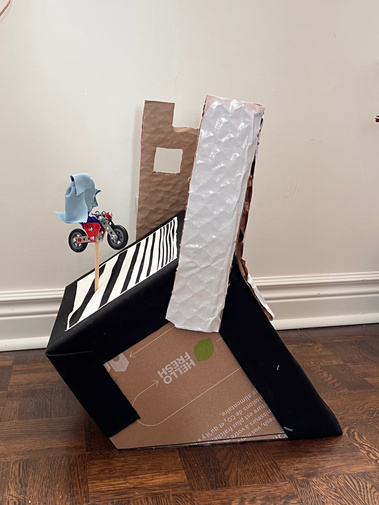 Side view of a cardboard box sitting at an angle, with cardboard buildings attached at the back. A wooden dowel holds up a metal toy motorcycle, and blue human shaped pipe cleaners sit in front the buildings