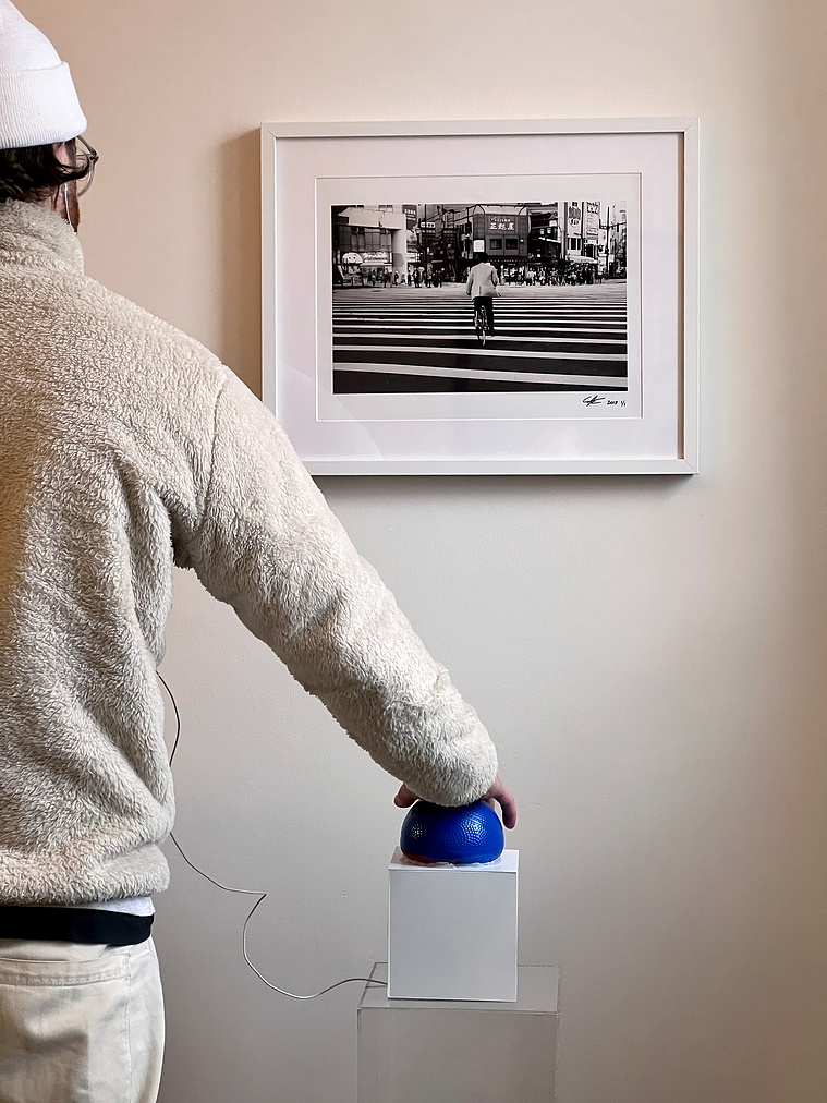 Black and white photograph hanging on a wall with a white plastic box with blue rubber ball on top sitting on a clear acrylic pedestal. A man wearing white to the left holding his right hand on the ball, while headphones go up to his ear