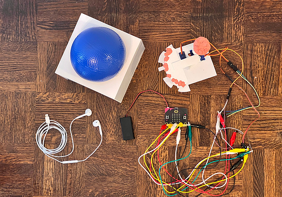 Overhead view of a white cube with blue ball on top, wrapped white headphones, a plastic tower with several wires coming out going to a Microbit and battery.
