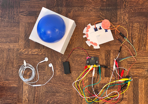 Overhead view of a white cube with blue ball on top, wrapped white headphones, a plastic tower with several wires coming out going to a Microbit and battery.