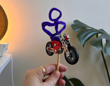 A metal toy motorcycle with a pipe cleaner shaped as a human sitting on top of a wooden dowel 