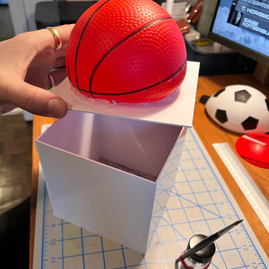 Half of a rubber toy basketball glued to the top of a white plastic box, with the lid open.