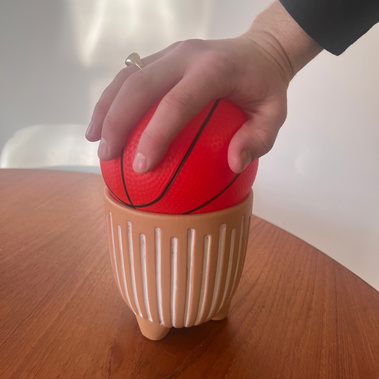 A rubber toy basketball sitting in a plant pot with a hand resting on top.