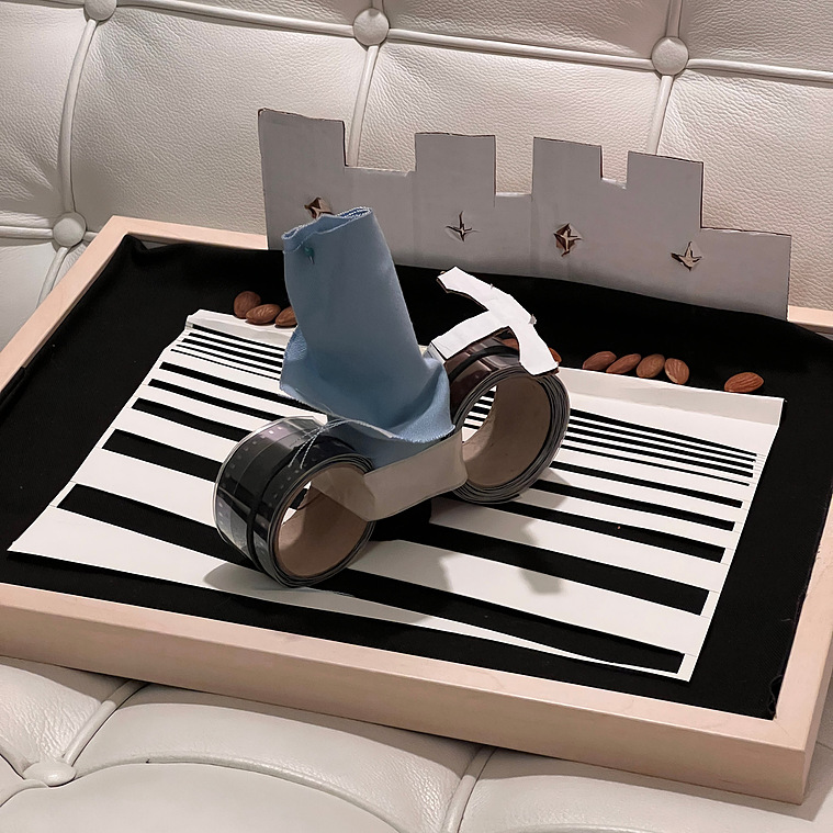 Picture frame laying flat with handmade bicycle made from rolls of film, and canvas, placed on top of a black canvas with white stripes. A row of almonds run across the back with cut cardboard in the shape of buildings