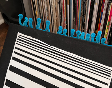 A cardboard box covered in black canvas with white paper stripes glued to it. A row of blue human shaped pipe cleaners are attached at the top.