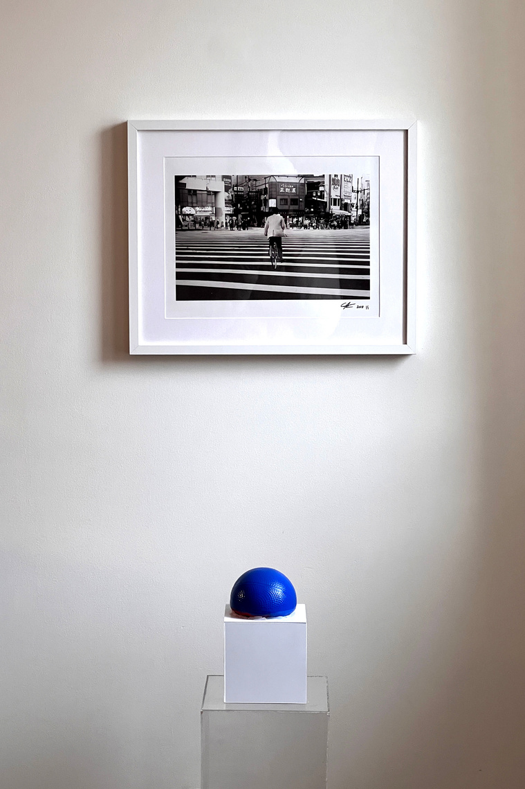 Black and white photograph hanging on a wall with a white plastic box with blue rubber ball on top sitting on a clear acrylic pedestal 