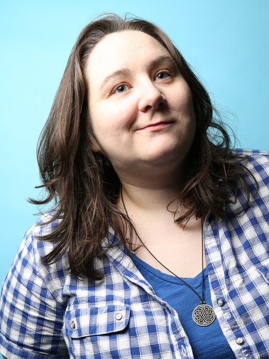 A portrait of Alanna McKibbon standing in a blue shirt with a blue and white plaid shirt layered over top. She has shoulder length wavy brown hair, blue eyes, is wearing a Celtic knotted necklace, and is smiling while looking up to the top right. 