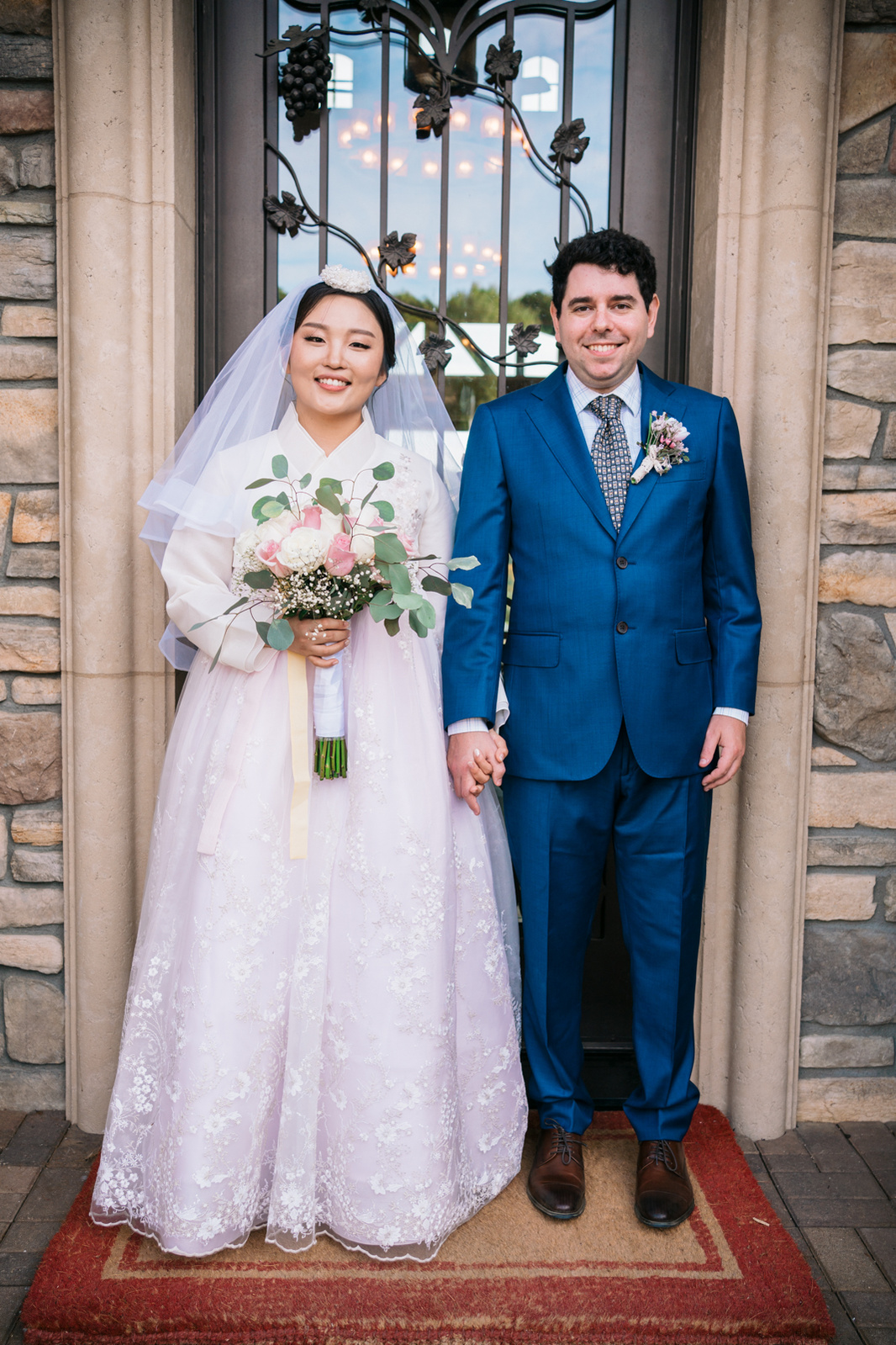 korean bride in her hanbok and jewish groom stand side by side hand in hand in front of a door