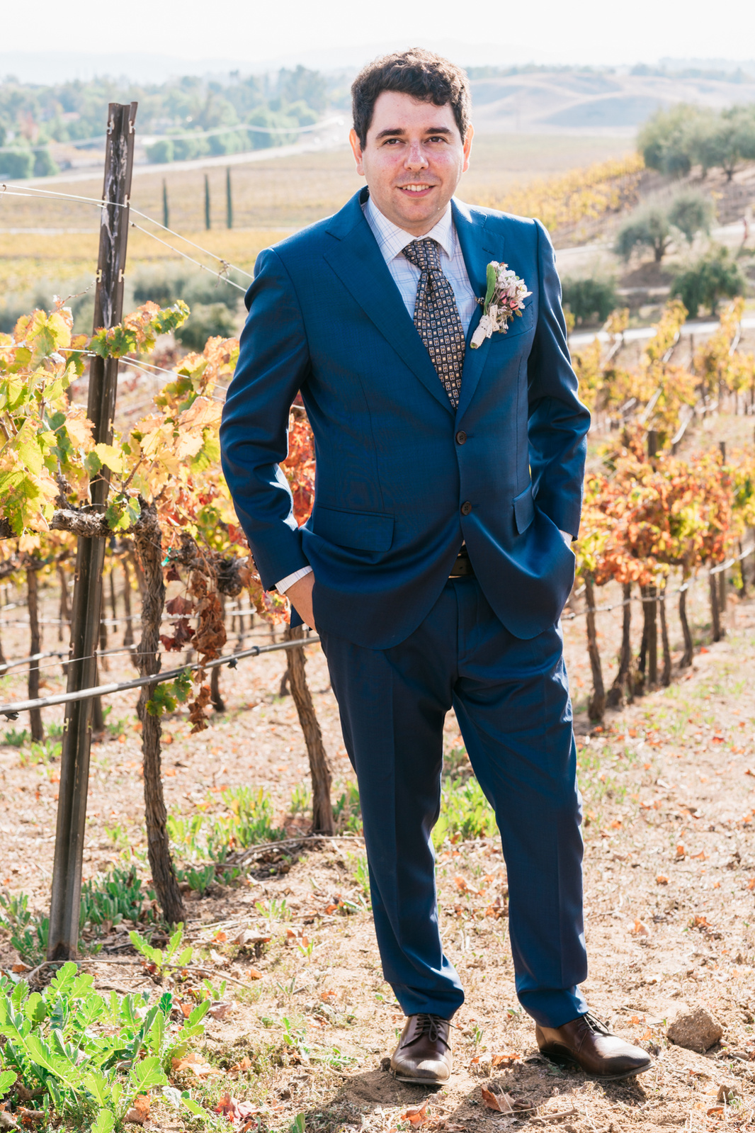 groom  hands in pockets poses among the vines in the vineyard