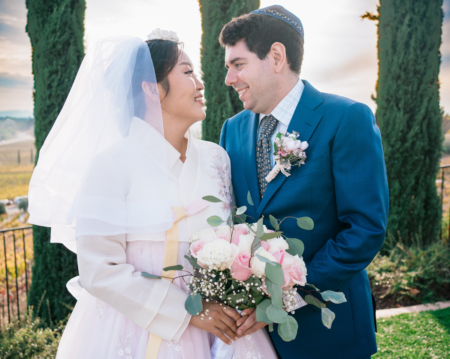 korean bride and jewish groom pose in front of cypress trees face to face hand in hand